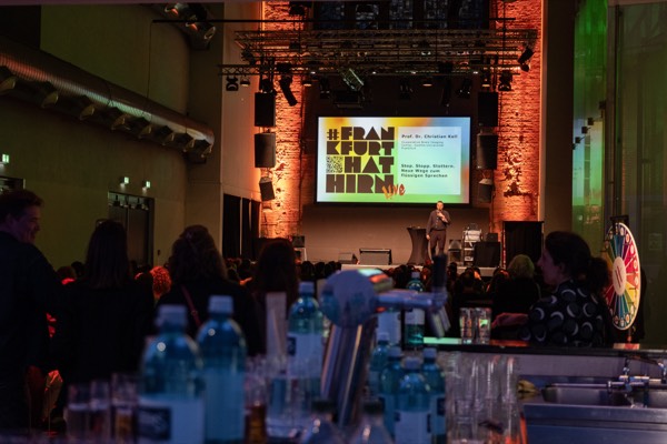 Blick über das Publikum auf die Bühne, auf der Leinwand das Logo von #Frankfurt hat Hirn Live und der Titel eines Flash Talks.