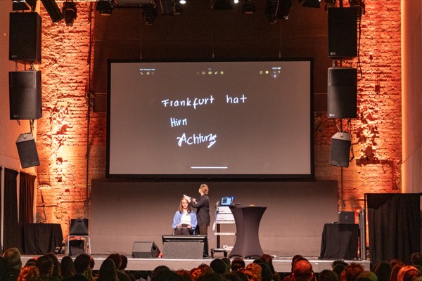 Zwei Personen auf der Bühne vor einer Leinwand mit dem handgeschriebenen Schriftzug „Frankfurt hat Hirn“, davor der volle Saal.