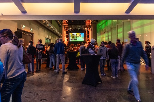 Volles Foyer mit Stehtischen, im Hintergrund die Leinwand mit dem Veranstaltungslogo.