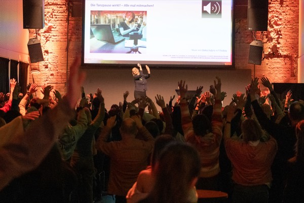 Das Publikum hebt bei der Mitmach-Tanzpause die Arme, auf der Leinwand steht „Die Tanzpause wirkt! – Alle mal mitmachen!“.